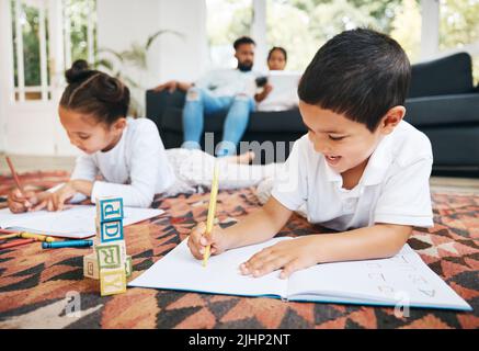 Little boy and girl drawing with colouring pencils lying on living room floor with their parents relaxing on couch. Little children sister and brother Stock Photo
