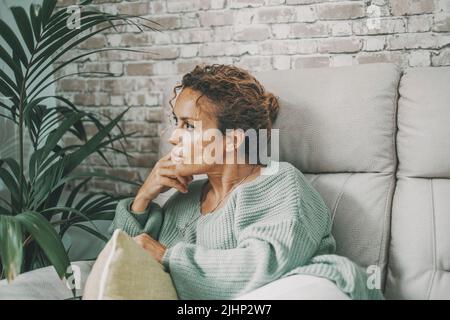 Thoughtful and worried lady at home sitting on the sofa looking serious and thinking a lot. Female portrait indoor. Lady sit on the couch touching his Stock Photo