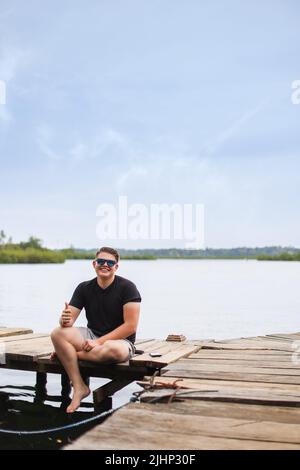 Young caucasian man sitting on pier with his thumb up smiling looking camera at bocas del toro panama Stock Photo