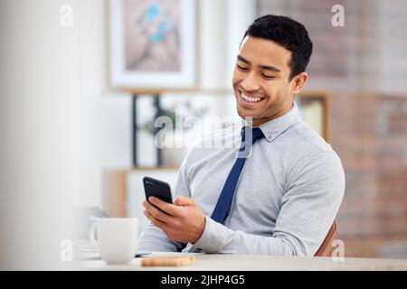 Young happy mixed race businessman holding and using a phone sitting in an office at work. One hispanic male businessperson smiling while using social Stock Photo