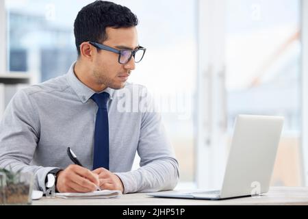 Focused mixed race businessman writing in a diary while working on a laptop alone in an office at work. One serious hispanic male businessperson Stock Photo