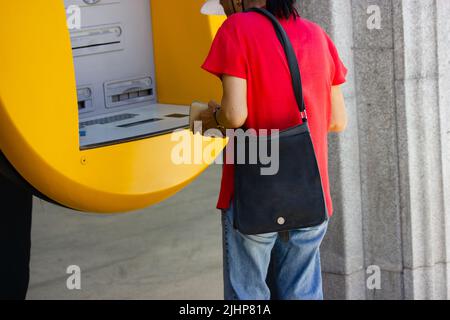Asian woman withdraws cash from yellow ATM. Currency, financial transactions. Chinese old lady withdrawing money outdoors on city street. Inserting cr Stock Photo