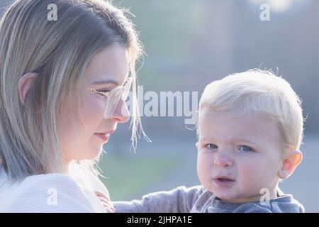 Happy young mother with a small child or toddler in her arms, hugging and cuddling, standing in the middle of a park in the rays of the rising morning sun. High quality photo Stock Photo