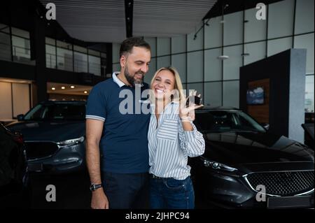 Happy caucasian couple hugging and woman holding new car keys in car showroom. Stock Photo