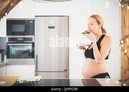 Beautiful sporty fit young pregnant woman having a healthy snack in home kitchen. Healty lifestyle concept. Stock Photo