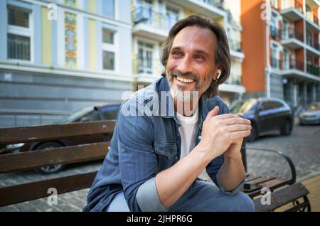 Smiling positive emotion middle aged man leaned his hands on knee looking sideways sitting at the bench in old town. Happy mature grey bearded man with positive emotions on his face. Stock Photo