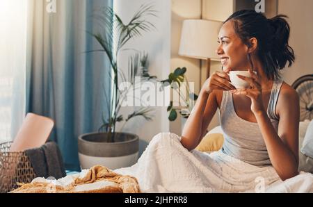A beautiful young Hispanic woman enjoying a warm cup of coffee for breakfast. One mixed race female drinking tea while sitting in bed and daydreaming Stock Photo