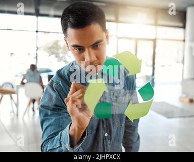 Young focused mixed race businessman drawing a recycle symbol on a glass window in an office at work. One serious hispanic businessperson designing a Stock Photo