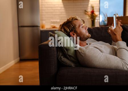 Ginger bristle man using mobile phone while lying on sofa at home Stock Photo