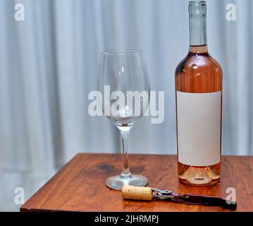 bottle of rosé wine with blank label with glass and a corkscrew on wooden table with white curtain in the background. Horizontal. pink wine tasting Stock Photo
