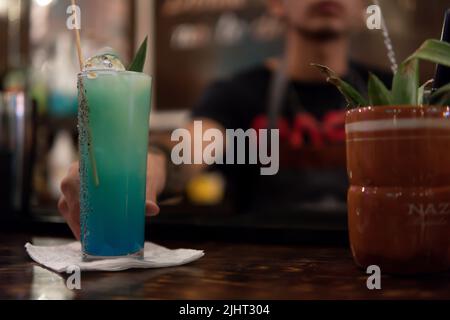 Blue alcoholic drink in the foreground being served by the waiter in the background. Drink on a bar counter with a pot next to it. Waiter serving a drink. Stock Photo