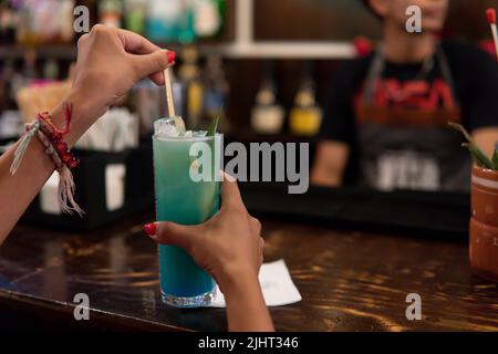 Hands of a girl mixing her alcoholic drink at a bar counter next to the bartender. Blue drink in the hands of a girl with bracelets. Counter of a bar with a waiter and a client. Stock Photo