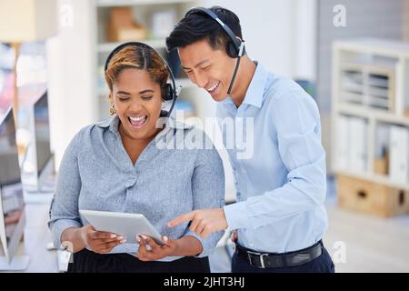 Two happy young diverse call centre telemarketing agents working on a digital tablet in an office. Friendly colleagues troubleshooting solution for Stock Photo