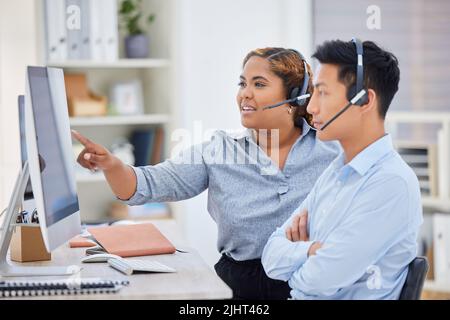 Young african american call centre telemarketing agent training new asian assistant on a computer in an office. Team leader troubleshooting solution Stock Photo
