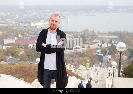 A young man with a red beard smiles with his arms folded on his chest against the backdrop of an autumn landscape. Stock Photo
