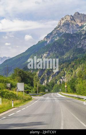 Beautiful road to the Alps in Austria leading to wooded slopes against a blue sky with clouds. Stock Photo