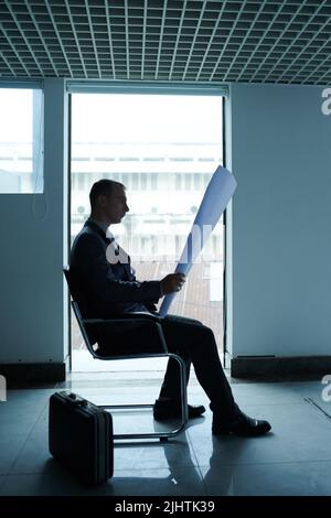 Side view of serious engineer sitting on chair and looking at big blueprint in his hands Stock Photo