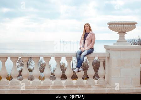 An adult woman with long blond hair sits on a parapet against the backdrop of the Crimean bridge. Stock Photo