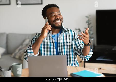 Happy Black Man Talking On Cellphone Using Laptop At Home Stock Photo