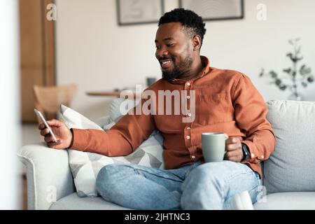 Happy Black Man Using Application On Smartphone Drinking Coffee Indoors Stock Photo