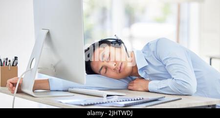 Close up of young asian businessman feeling tired and sleeping on the desk of a call center in a office. Male wearing wireless headset suffering from Stock Photo