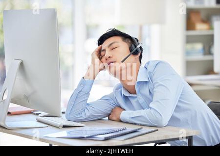 Close up of young asian businessman feeling tired and sleeping on the desk of a call center in a office. Male wearing wireless headset suffering from Stock Photo