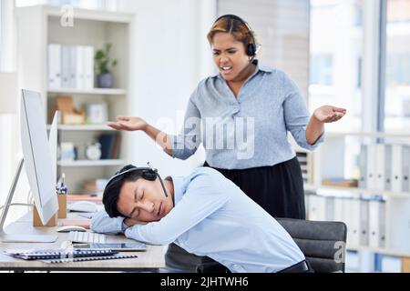 Close up of young asian businessman feeling tired and sleeping on the desk of a call center in a office while his hispanic female colleague looks Stock Photo