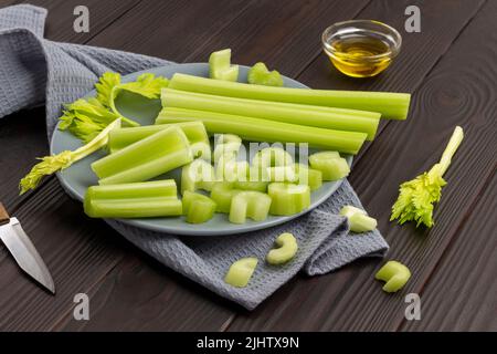 Chopped celery stalks on gray plate on napkin. Knife and olive oil in bowl. Top view. Wooden background. Stock Photo