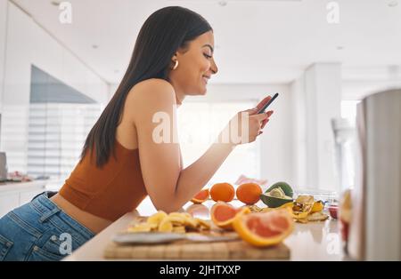 Im making breakfast, what about you. a woman using her cellphone while making a smoothie at home. Stock Photo