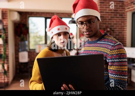 Multiethnic team of colleagues working on laptop during winter season in office with christmas tree and decorations. Man and woman doing teamwork, celebrating festive holiday with ornaments. Stock Photo