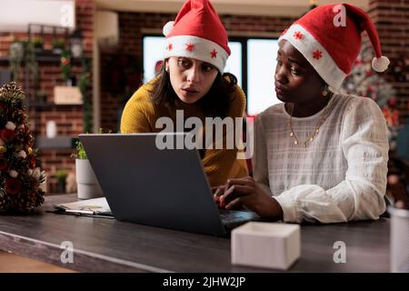 Multiethnic team of women with santa hat working on business at laptop, sitting in festive office filled with christmas decorations and ornaments. Doing teamwork and celebrating holiday. Stock Photo