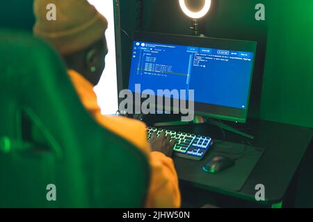 Concentrated bearded black guy in an orange outfit looking away, sitting on a gaming swivel chair in a dark room with neon lights on walls. High quality photo Stock Photo
