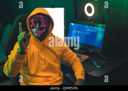 Black man wearing a dark mask with pink patterns covering his face, pointing his finger at the camera, sitting in front of a big modern computer screen. High quality photo Stock Photo