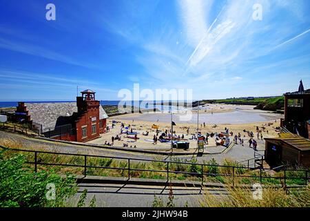 Cullercoats Bay beach wideangle vista enjoying the sunshine with canoeists surfers lifeguards tents RNLI station and the Dove Marine Lab deep blue sky Stock Photo