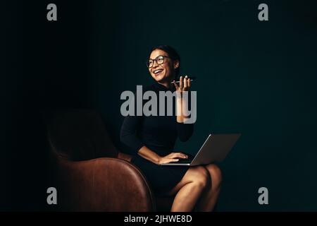 Asian female sitting in chair with laptop and talking on mobile phone. Happy businesswoman talking on cellphone and looking away. Stock Photo