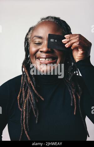 Middle-aged woman with dreadlocks smiling cheerfully while holding a credit card in front of her eye. Happy mature woman recommending electronic banki Stock Photo