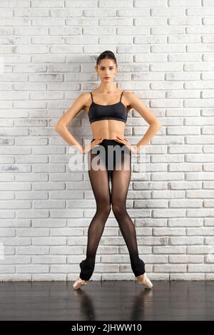 Full body of fit female ballet dancer in black bra and pointe shoes looking at camera against white brick wall in studio Stock Photo