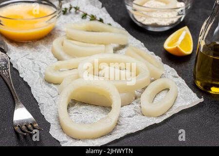 Squid cut into rings on paper.  Fork and oil in bottle. Flour and beaten raw egg in bowls. Top view. Black background Stock Photo