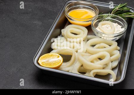 Squid cut into rings in baking tray. Flour and beaten raw egg in bowls. Top view. Copy space. Black background Stock Photo