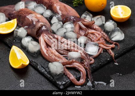 Fresh squid with tentacles with ice and lemon on stone board. Close up. Top view. Black background Stock Photo