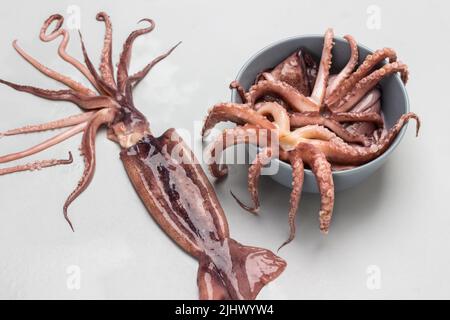 Fresh squid with tentacles in bowl and on gray plate. Flat lay. Gray background. Stock Photo