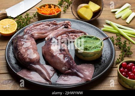 Raw squid with tentacles and bowl of seaweed on black plate. Carrots, potatoes, knife and celery stalks on table. Top view. Wooden background. Stock Photo