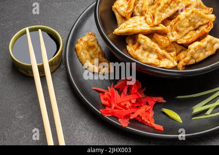 Fried gyoza dumplings in sauce on black plate. Soy sauce in bowl, chopsticks. Top view. Black background Stock Photo