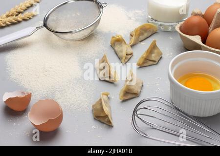 Raw gyoza, flour and sieve on table. Broken egg in bowl, whisk and eggshell. Top view. Gray background. Stock Photo