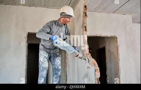Man builder in construction helmet drilling wall with hammer drill. Male worker using drill breaker while destroying wall in apartment under renovation. Demolition work and home renovation concept. Stock Photo