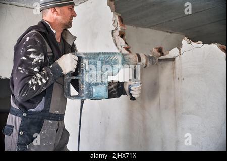 Man builder in workwear drilling wall with electric hammer drill. Male worker using electric drill while breaking wall in apartment under renovation. Demolishing and construction destroying concept. Stock Photo