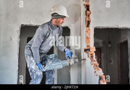 Side view male professional of demolition walls in construction helmet using perforator in mode of jackhammer and destroying interroom brick septum. Redevelopment, reconstruction of housing. Stock Photo