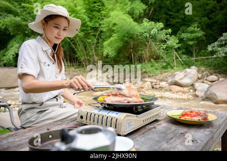 Pretty and charming young Asian female traveler preparing her picnic lunch, making a barbecue steak near the river. Stock Photo