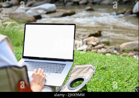 Close-up image, Female using notebook laptop computer while taking a vacation break at the natural campground. Laptop white screen mockup. Stock Photo