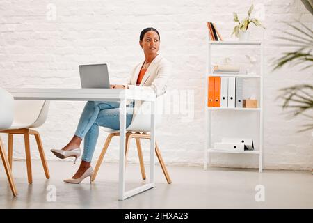 Young mixed race businesswoman thinking while working on a laptop at a table in an office at work. One hispanic businessperson typing on a laptop Stock Photo